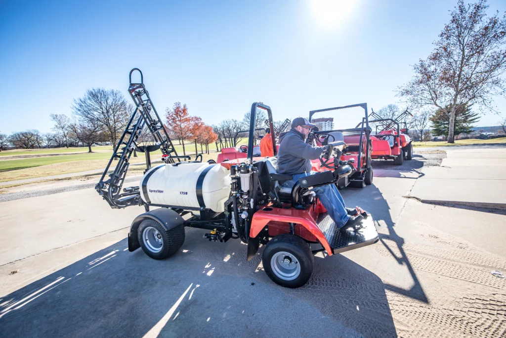 Golf course sprayer, Smithco Spray Star 1750, with operator, advanced spray boom, and hydrostatic drive, parked on pavement, showcasing precision turf maintenance equipment.