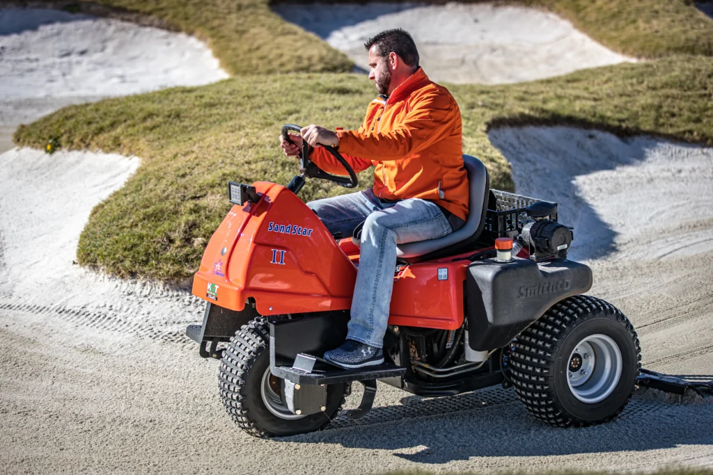 Man operating Smithco Sand Star II bunker rake on sand surface, designed for golf course maintenance, showcasing robust performance and precision in turf care.