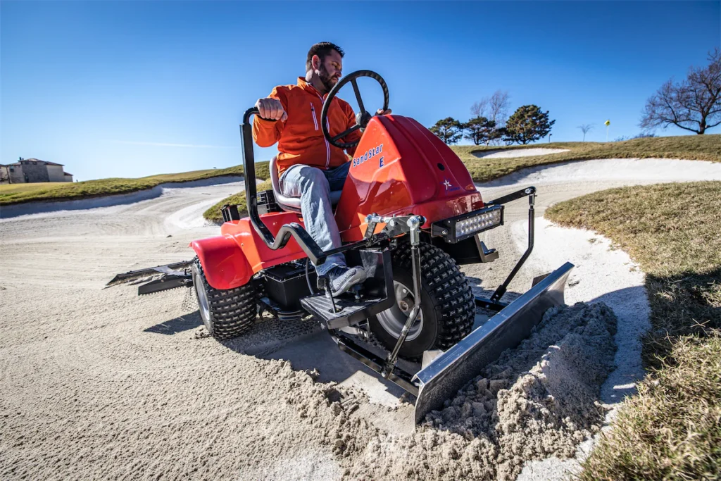 Man operating the Smithco Sand Star E bunker groomer on a golf course, showcasing energy-efficient performance and superior traction.