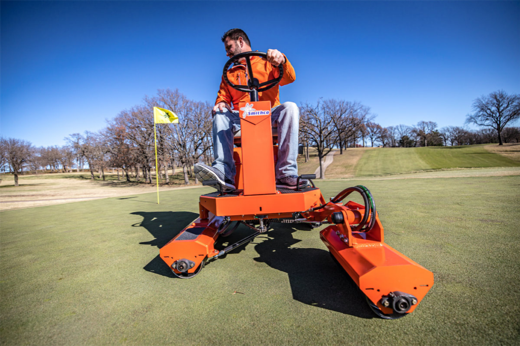 Man operating Smithco Tournament Ultra Wide XL 7000 greens roller on golf course, with flag in background and clear blue sky.